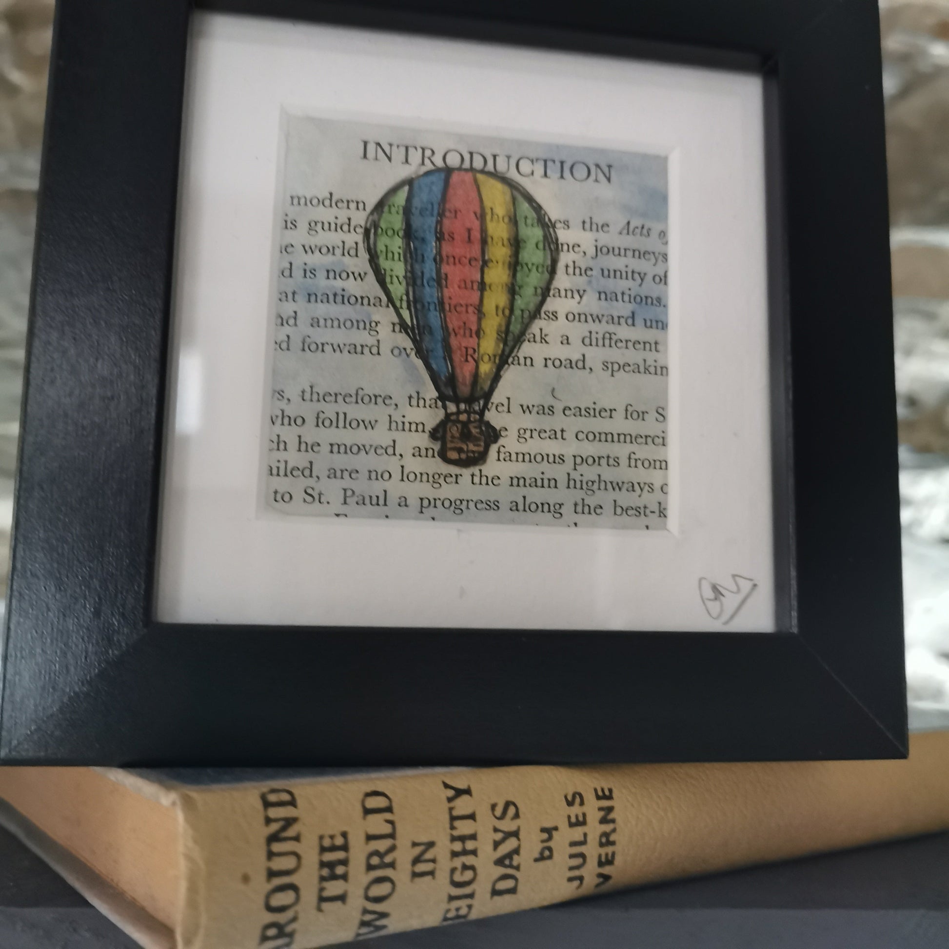 A freestanding black 12.5 cm box frame on a bookshelf with the book around the World in 80 days against a backlit slate wall. A 6cm internal frame with a book page and a hand-drawn watercolour illustration of a hot air balloon.