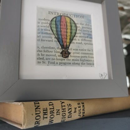 A freestanding grey 12.5 cm box frame on a bookshelf with the book around the World in 80 days against a backlit slate wall. A 6cm internal frame with a book page and a hand-drawn watercolour illustration of a hot air balloon.