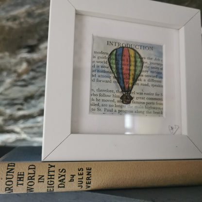 A freestanding white 12.5 cm box frame on a bookshelf with the book around the World in 80 days against a backlit slate wall. A 6cm internal frame with a book page and a hand-drawn watercolour illustration of a hot air balloon.