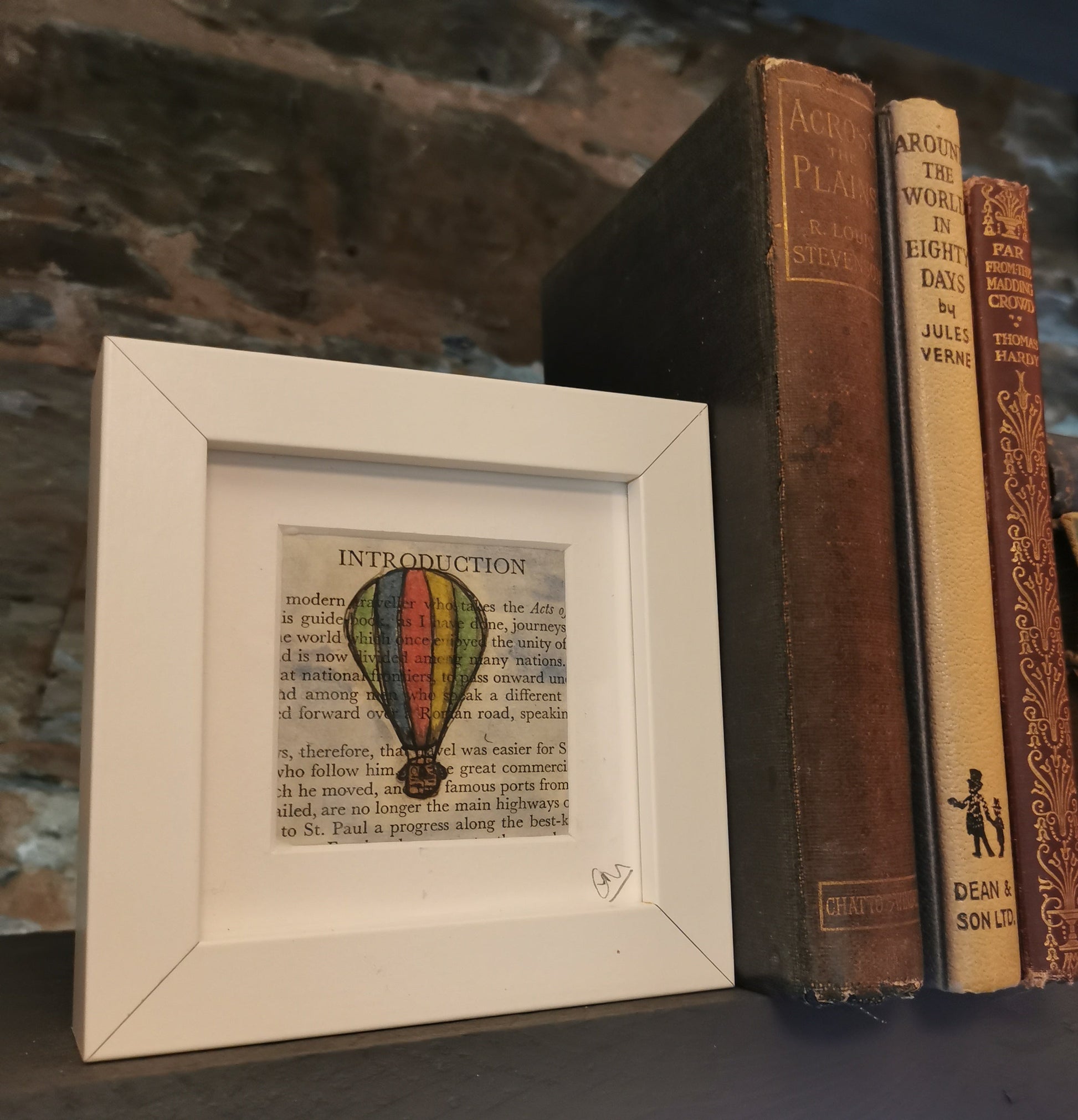 A freestanding white 12.5 cm box frame on a bookshelf with the book around the World in 80 days against a backlit slate wall. A 6cm internal frame with a book page and a hand-drawn watercolour illustration of a hot air balloon.