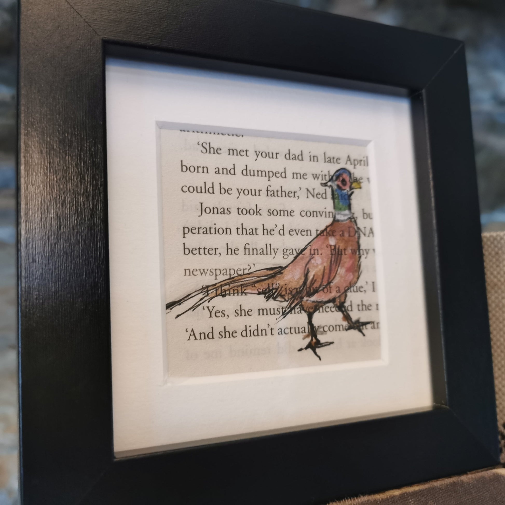 A freestanding black 12.5 cm box frame on a bookshelf against a backlit slate wall, with a 6cm internal frame with a book page and a hand-drawn watercolour illustration of a pheasant.